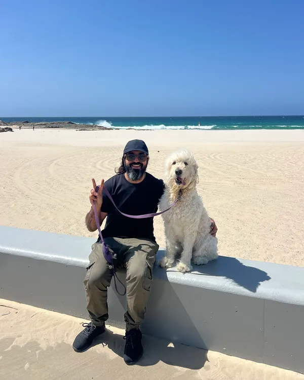 AJ Awan sitting on a sea wall with his dog, a white groodle, on a Gold Coast beach with the ocean behind them